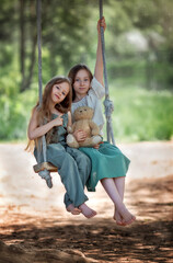 Happy laughing kids girls sisters with long hair enjoying a swing ride with a teddy bear toy on a sunny summer day