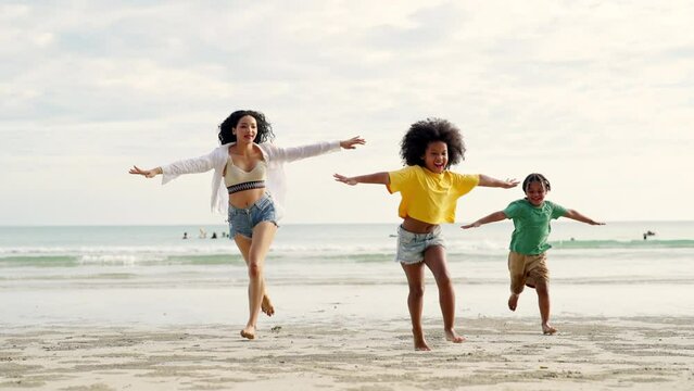Happy African family mother with little daughter and son running and playing sea water together on the beach at summer sunset. Mom with child boy and girl enjoy outdoor lifestyle vacation at the sea