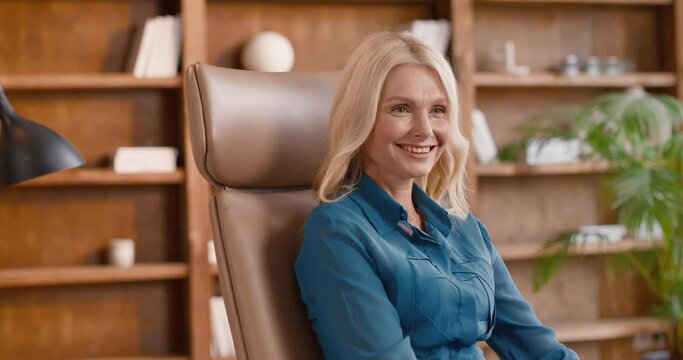 Elegant blond woman turning head and smiling sincerely while sitting in brown armchair with wooden shelves on background. Happy mature psychotherapist welcoming new patient at home office.