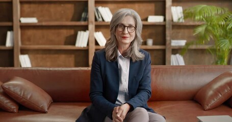 Pleasant grey haired woman sitting on comfy brown couch in bright office. Portrait of smiling skilled female psychologist wearing trendy glasses and formal suit.
