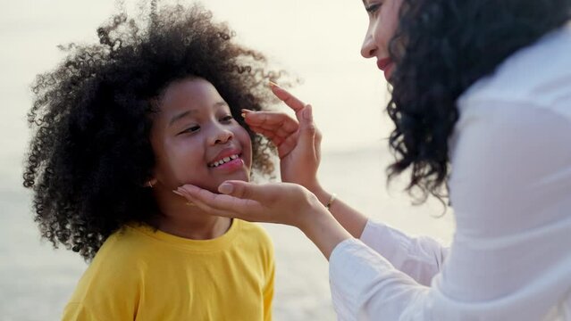 Happy African family mom and child girl kid enjoy and fun outdoor lifestyle on beach vacation. Mother applying sunblock to little daughter while walking together on tropical beach at summer sunset.
