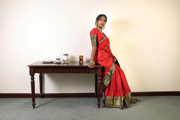 Indian woman wearing red orange saree jewellery choker set necklace jhumka earring maang tikka waist chain stand pose look see smile mood expression standing siting retro vintage table   