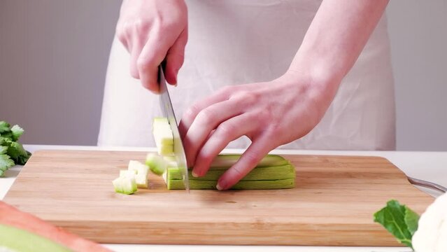Woman cuts courgette into cubes on a wooden cutting board on white table