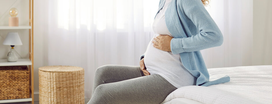 Woman expecting baby with love and tenderness strokes her pregnant tummy while sitting on bed. Cropped side view image of unknown pregnant woman in bedroom on bed on window background. Banner.