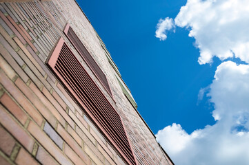 Fototapeta na wymiar Brick wall of a multi-storey building with ventilation grilles, against the background of a blue sky and white clouds. Modern urban architecture.