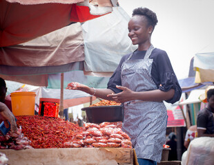 An African Nigerian female trader, seller, business woman or entrepreneur with an apron, showing her displayed goods to attract potential customers in a market