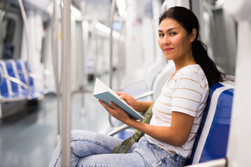 Oriental woman with book in hands sitting on bench in subway train and looking in camera.