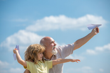 Young grandson and old grandfather with paper plane over blue sky and clouds. Men generation granddad and grandchild.
