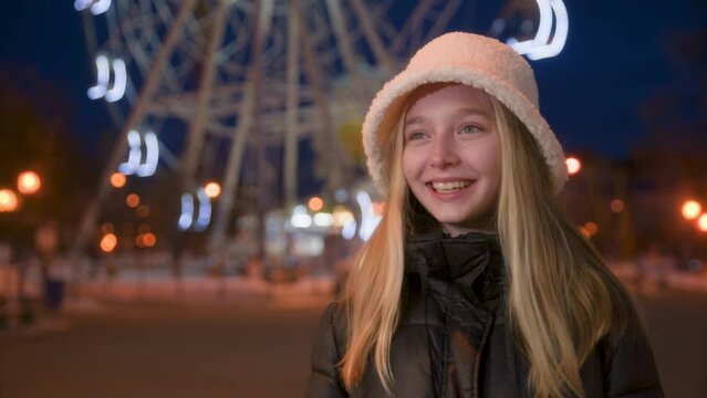 Cheerful teen blonde girl in white fur bucket hat laugh swaying and nodding by head against illuminated ferris wheel background in amusement park at night. Close-up