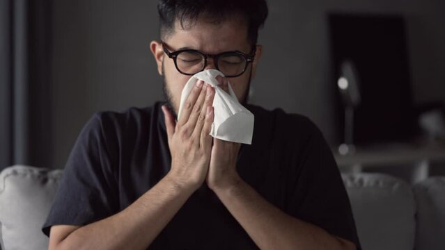 Asian man sick and sneeze with tissue paper on sofa at home. Male blowing nose suffering from cold. Cold and fever concept.