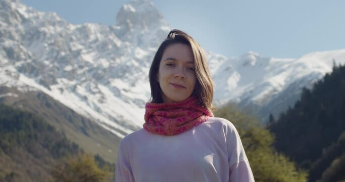 Portrait of young woman looking at camera and smiling on background of snowy mountains. Tourist girl standing outdoors alone enjoying beautiful moment in nature. Happy tourist during travel 