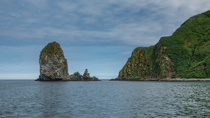 Bizarre rocky islands rise in the Pacific Ocean. Green vegetation on steep slopes. Clouds in the blue sky. Kamchatka.