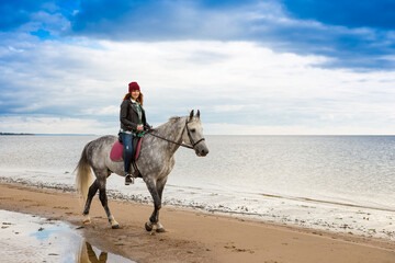 wearing jeans and jacket horsewoman is in the sabble on a gray horse along seacoast
