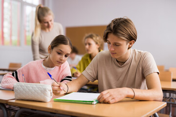 Teenager boy and younger girl sitting at desk in classroom and doing tasks.