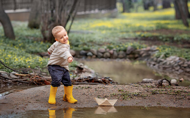Portrait of adorable little kid boy in rubber boots launch paper boats in a puddle in spring. Origami.
