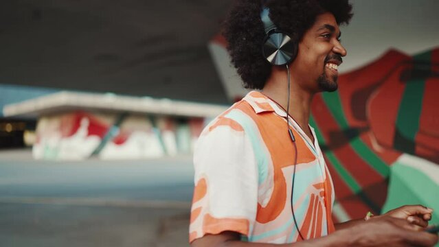 Close-up of cheerful young African American man wearing shirt listening to music in headphones and dancing on urban city background. Lifestyle concept. Slow motion
