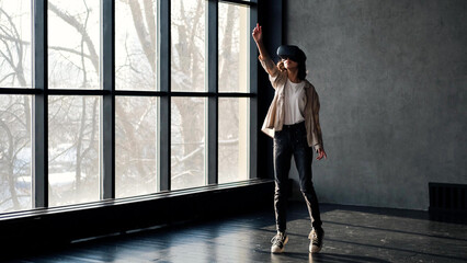 Fototapeta na wymiar A teenage girl in virtual reality glasses stands in a gray room near a large window.