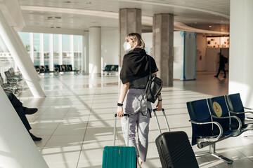 Fototapeta na wymiar Back view of woman wearing black T-shirt, grey trousers, disposable mask, going, holding turquoise, black suitcases.