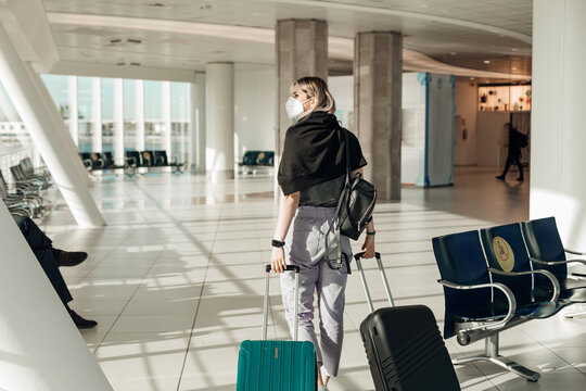 Back view of woman wearing black T-shirt, grey trousers, disposable mask, going, holding turquoise, black suitcases.