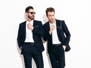 Portrait of two handsome confident stylish hipster lambersexual models. Sexy modern men dressed in black elegant suit. Fashion male posing in studio near white wall in sunglasses