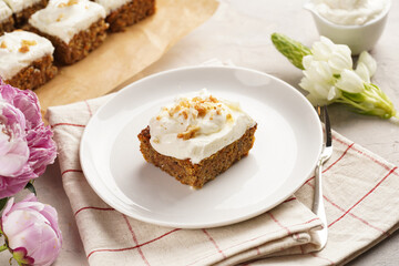 Homemade pastry carrot-walnut cake with grounded almonds and hazelnuts and white cream cheese top layer on white plate on pink checkered napkin