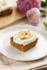 Homemade pastry carrot-walnut cake with grounded almonds and hazelnuts and white cream cheese top layer on white plate on pink checkered napkin