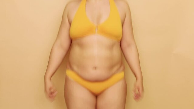 Unrecognizable fat plump overweight woman wearing yellow bra, bikini, jumping, showing excess flabby subcutaneous fat of belly on beige background. Body positive, massage, weight loss, liposuction.