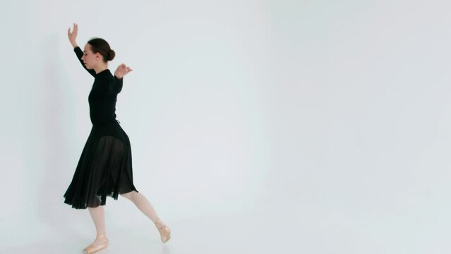 A ballerina in black clothes performs a classical dance in a white studio and demonstrates a graceful jump. Slow motion