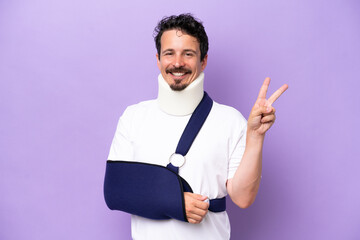 Young caucasian man wearing a sling and neck brace isolated on purple background smiling and...