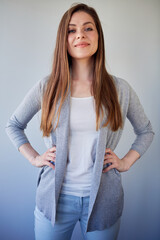 Smiling woman with long brown hair keeps hands on hip. Isolated portrait of girl wearing casual gray clothes.