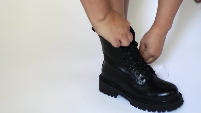 Cropped photo of woman standing in white socks, putting on black glossy boots with shoelaces, zipping up on white background. Advertisement, unisexual fashion, shoes for women. Close-up. Studio shot.