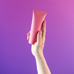  Mockup of a pink tube with a cosmetic product in a female hand on a purple background. Copy space. Banner.