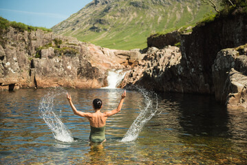 A woman wearing a green swimming suit splashes water with both hands while bathing in the River Etive in Glen Etive, Scottish Highlands, UK, with a waterfall and a mountain in the background.