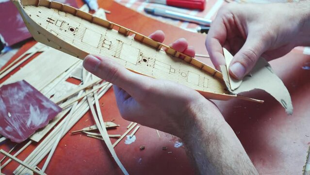 Hands of man adjusts plywood details for ship model, grinding on sandpaper. Process of building toy ship, hobby and handicraft. Table with various materials, parts and devices for work. 4k video