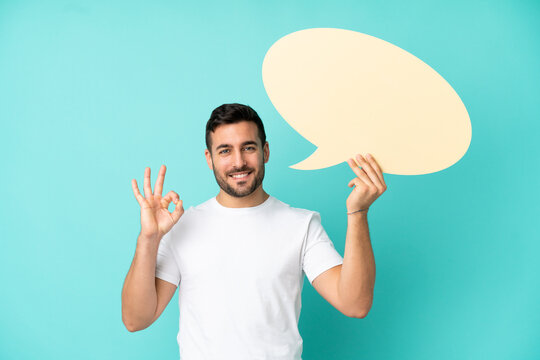 Young handsome caucasian man isolated on blue background holding an empty speech bubble and doing OK sign