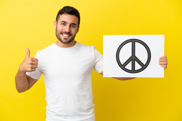 Young handsome caucasian man isolated on yellow background holding a placard with peace symbol with...
