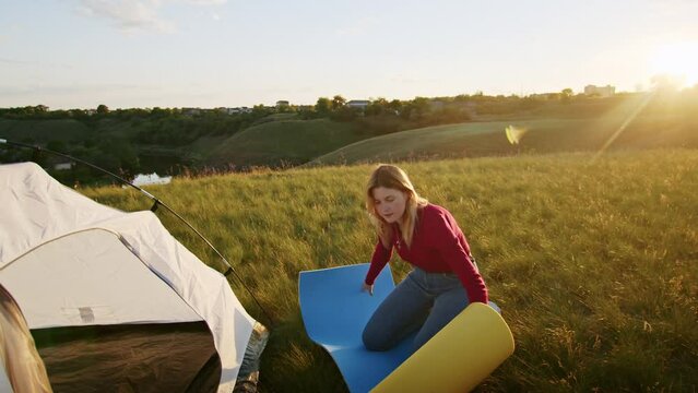 a young woman happily set up a tent on the mountain hills during sunset, for rest after a hike. a woman lays out a karimat. rest of one person in nature, in the mountains.