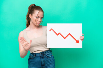 Young caucasian woman isolated on green background holding a sign with a decreasing statistics arrow symbol and doing stop sign