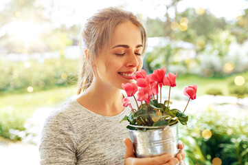 farming, gardening and people concept - happy young woman smelling pink cyclamen flowers in pot at summer garden - obrazy, fototapety, plakaty