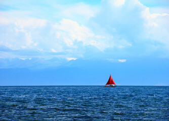 Beautiful romantic view of lonely sailing ship with red sheet against the background of distant mountain range and cloudy blue sky, Issyk-Kul Lake, Kyrgyzstan, Central Asia