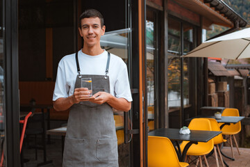 Young pleasant man cafe worker in apron serving table in local stylish coffeehouse or coffee shop