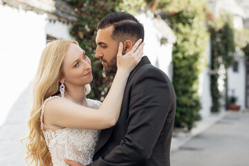 Naklejka premium Portrait of young blonde bride in white dress and brunet groom in suit embracing and looking at each other on city street in Italy, urban background. Beautiful and romantic wedding, happy couple