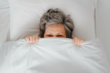 Concept depression, laziness, frustration, female mental health. Tired senior woman with gray hair covered with blanket and looking at camera while lying on bed in bedroom, top view