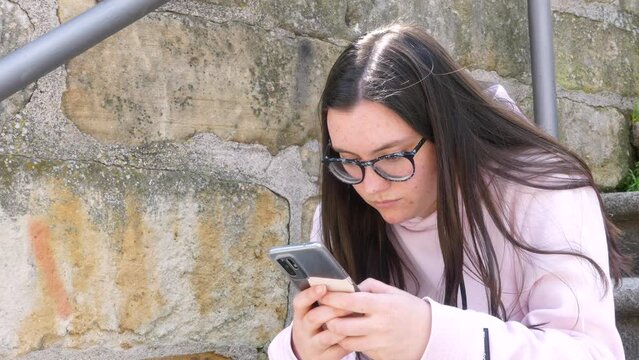 Cute nerd girl with glasses and dressed in pink chats using her smart phone on the street, outdoors video.