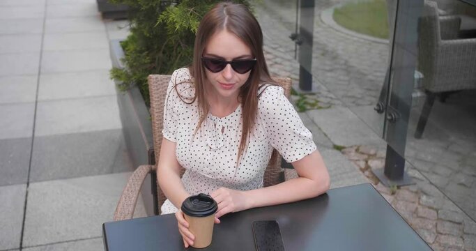 Beautiful young woman drinking tasty coffee and receiving call on mobile phone at outdoor cafeteria