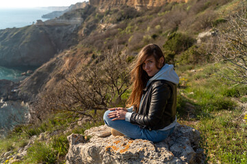 Woman tourist enjoying the sunset over the sea mountain landscape. Sits outdoors on a rock above the sea. She is wearing jeans, a blue hoodie and a black leather jacket. Healthy lifestyle, harmony and