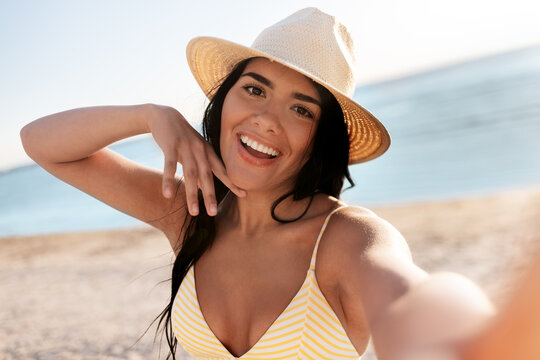 people, summer and swimwear concept - happy smiling young woman in bikini swimsuit and straw hat taking selfie on beach