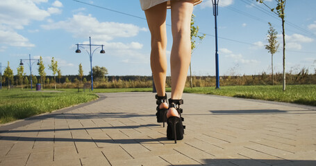 camera follows long female legs in high heels. A woman walks alone along the pavement in a park...
