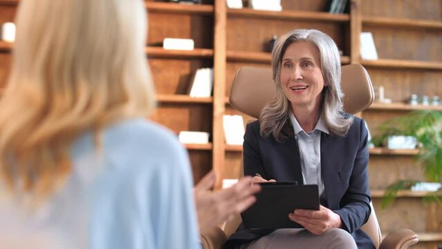 Attentive female psychologist in formal wear sitting with clipboard in cabinet and listening caucasian woman with blond hair during therapy session. Concept of mental health and work with emotions.