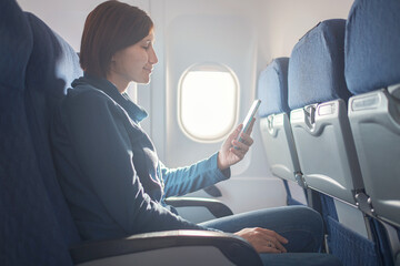 Young beautiful woman sitting at window of plane during the flight. reading a book on a smartphone,...